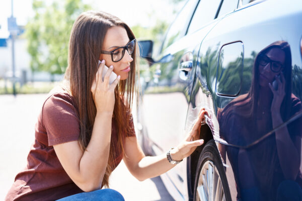 Woman on the phone inspecting a scratch above the back left tire of her car.