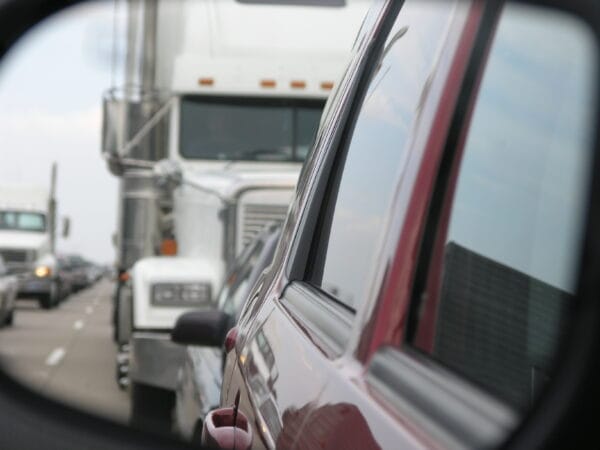 A white semi-truck closely follows a red car, seen through the car’s side mirror.