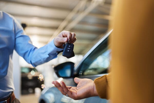 A person handing over car keys to another individual in front of a parked vehicle.