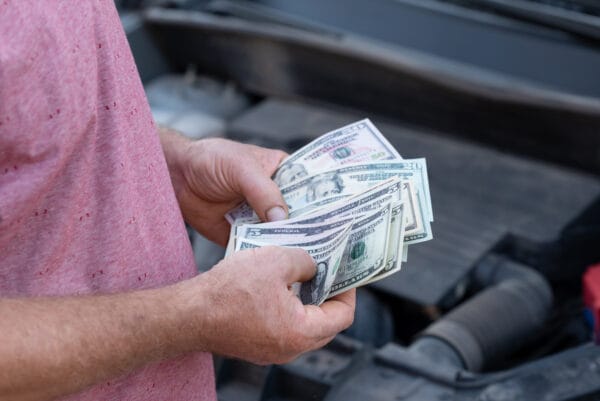 Close-up of a person in a red shirt holding a stack of U.S. dollar bills near a vehicle engine.