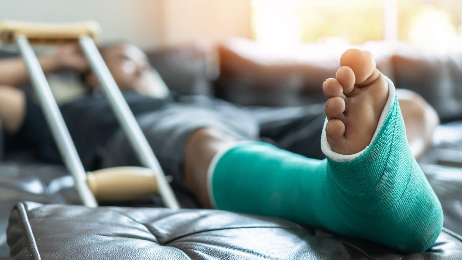 A person with their leg and foot in a green cast lays on the couch with one of their crutches next to them.