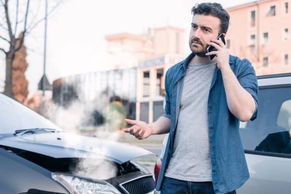 An upset man on the phone, standing next to a car that is badly damaged and has smoke coming out.