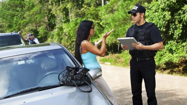 A woman is leaning against a car with her purse on the car next to her while she talks to a police officer. The police officer is holding a clipboard.