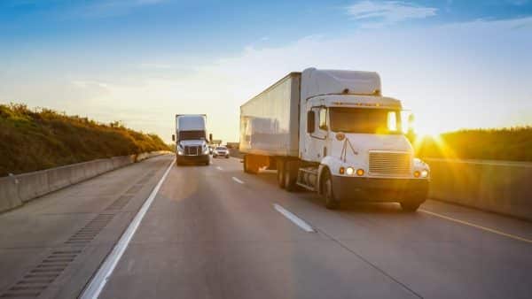 Two semi-trucks driving on a highway at sunset, with cars following behind.