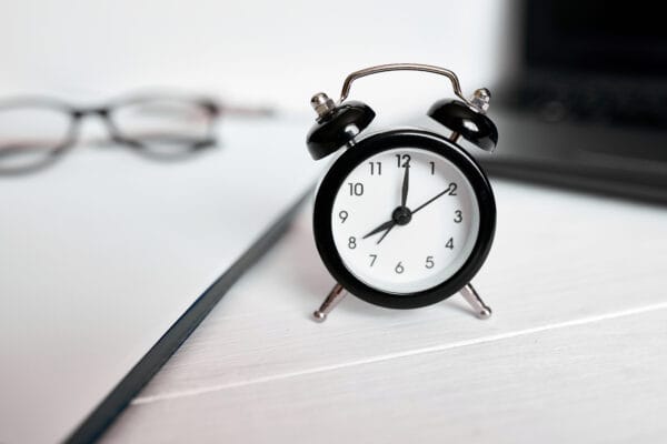 A small black alarm clock on a desk, with a notebook, glasses, and a laptop in the background.